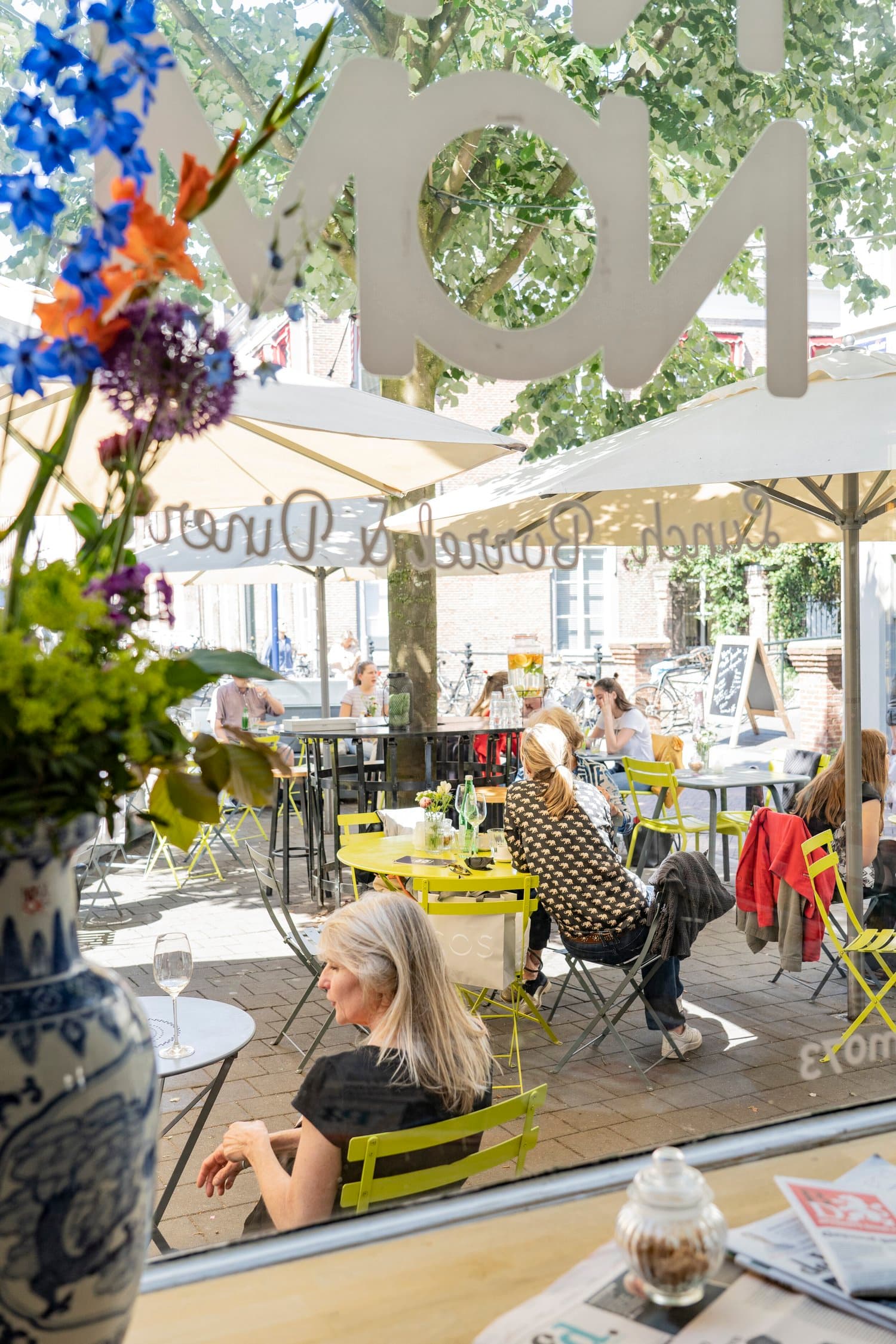 Doorkijk door het raam met NOM NOM-belettering naar het zonnige terras met bloemen op de vensterbank.