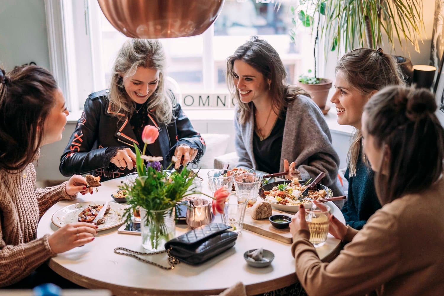 Vijf gasten aan een ronde tafel binnen bij NomNom, met bowls, brood en wijn onder een koperen hanglamp.