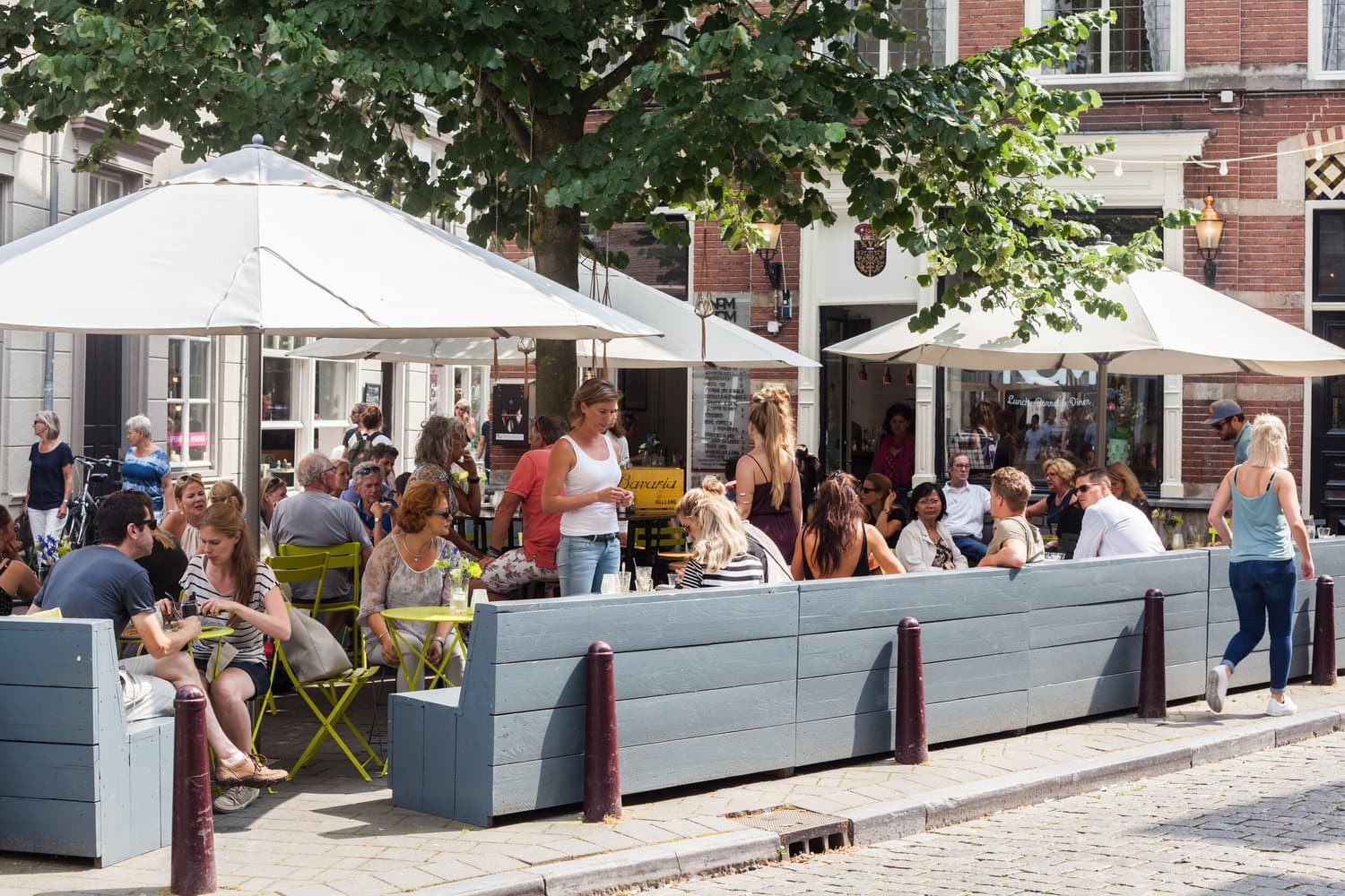 Het volle terras van NomNom op een zonnige dag, met witte parasols, limoengroene stoeltjes en een grote boom voor de historische gevels.